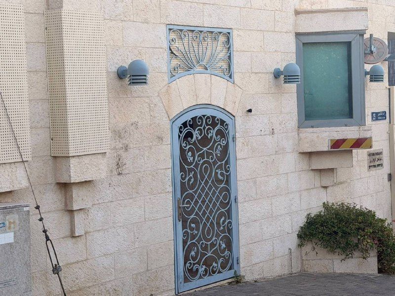 דלת מתכת מעוטרת בחזית מבנה אבן, לצד חלון ומנורות קיר.