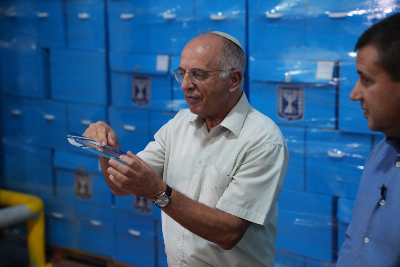 גבר חובש כיפה מחזיק חפץ כחול מול ערימות ארגזים כחולים, ולצדו גבר נוסף.