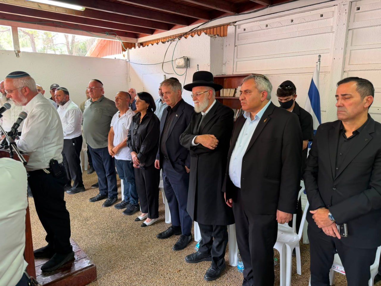 גברים ואישה עומדים בשורה באולם לצד מיקרופונים ודגל ישראל.