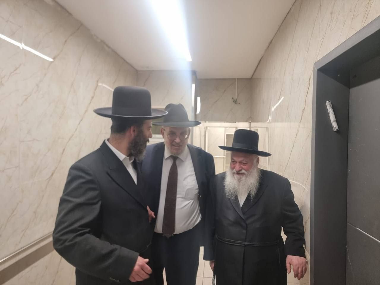שלושה גברים בחליפות כהות ובכובעים שחורים עומדים במסדרון ליד מעלית.