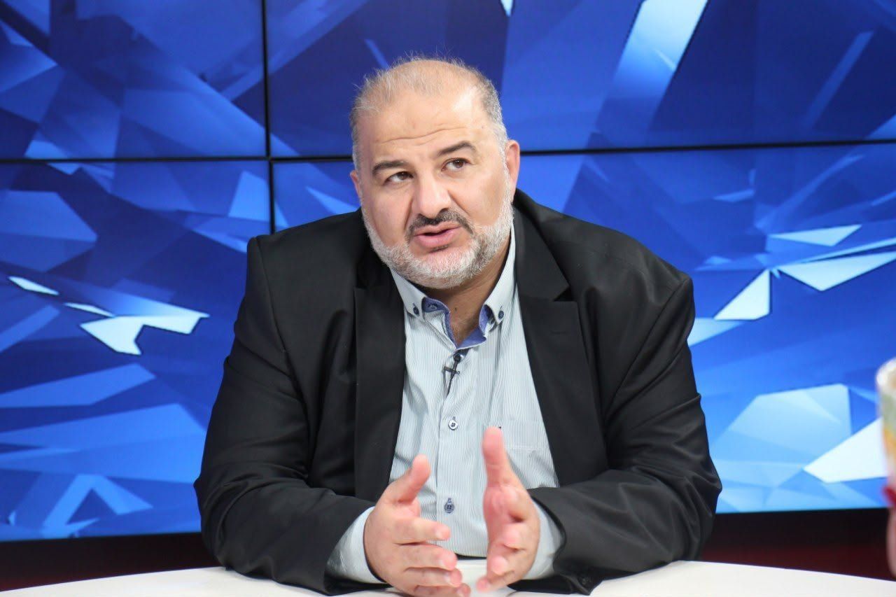מנסור עבאס יושב ליד שולחן ומחווה בידיו על רקע מסכים כחולים.