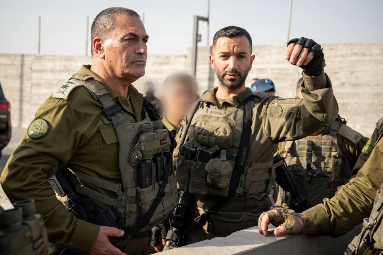 קצינים וחיילים במדים ובאפודי מגן עומדים בחוץ, ואחד מהם מרים יד ומסמן.